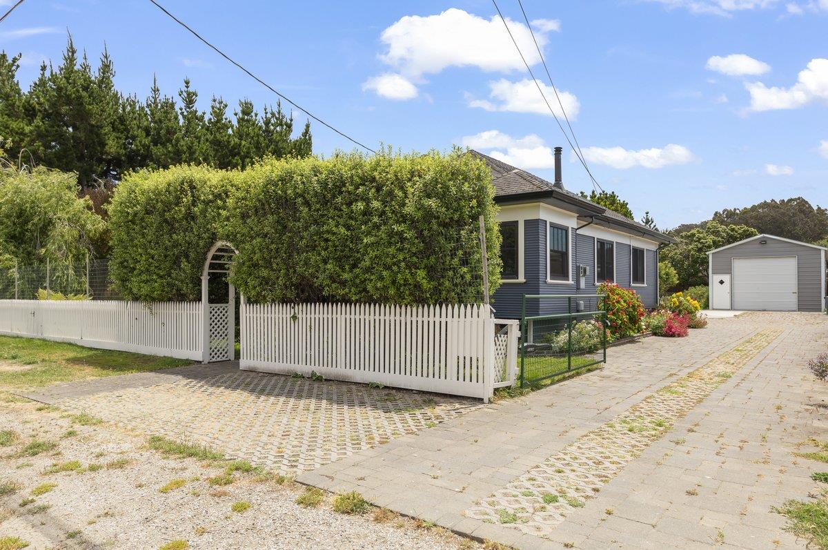 Gray home behind a tall hedge and white picket fence, with a paved driveway and detached garage.