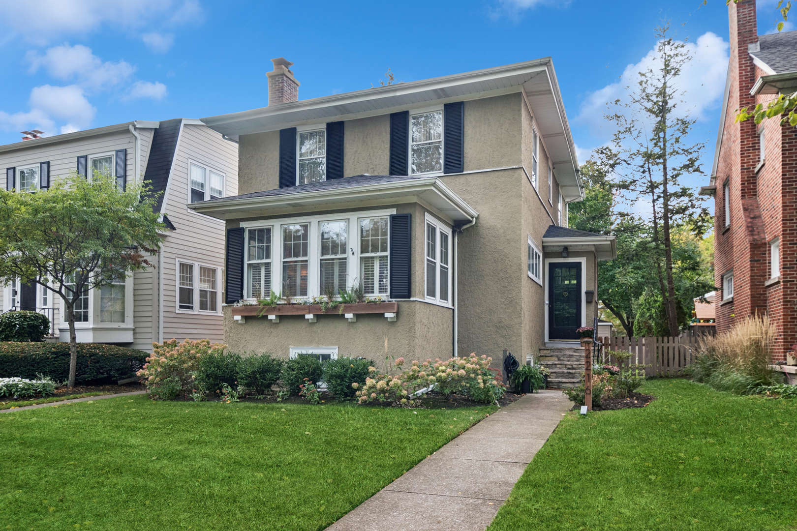 Two-story stucco house with dark shutters, a bay window, flower beds, and a walkway to the front door.