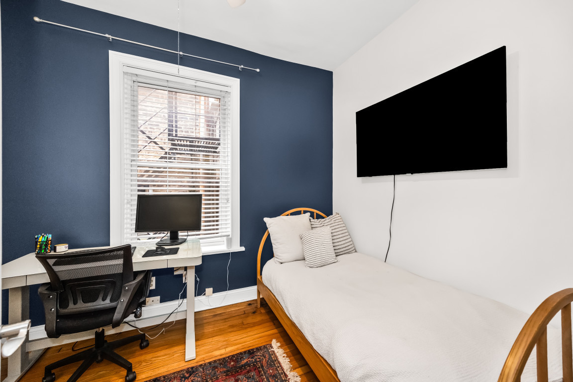 Bedroom with a single bed, wall-mounted TV, and computer desk beside a window.