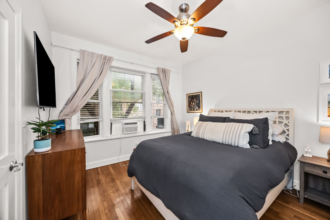 Bedroom with a large gray-covered bed, geometric headboard, hardwood floors, and curtained windows with a window air conditioner.