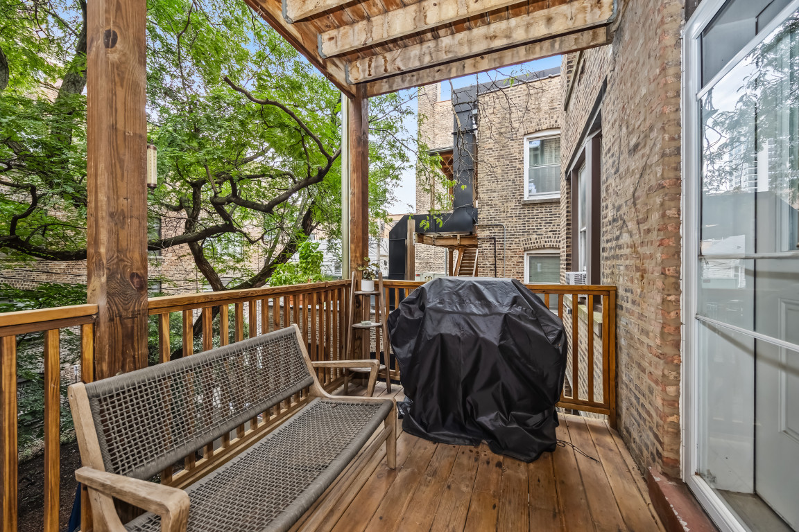 Wooden deck balcony with a woven bench, covered grill, and leafy trees beyond the railing.