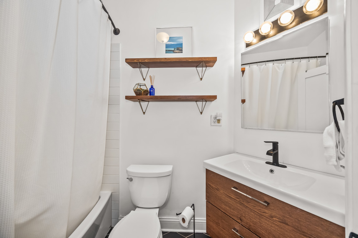 Bathroom with wood-front vanity, large mirror, floating shelves, toilet, and curtained bathtub.