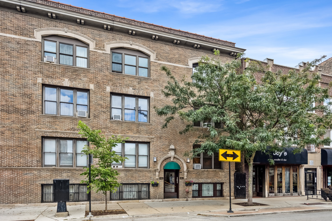 Brick residential building with arched windows, a central entrance, and trees along the sidewalk.
