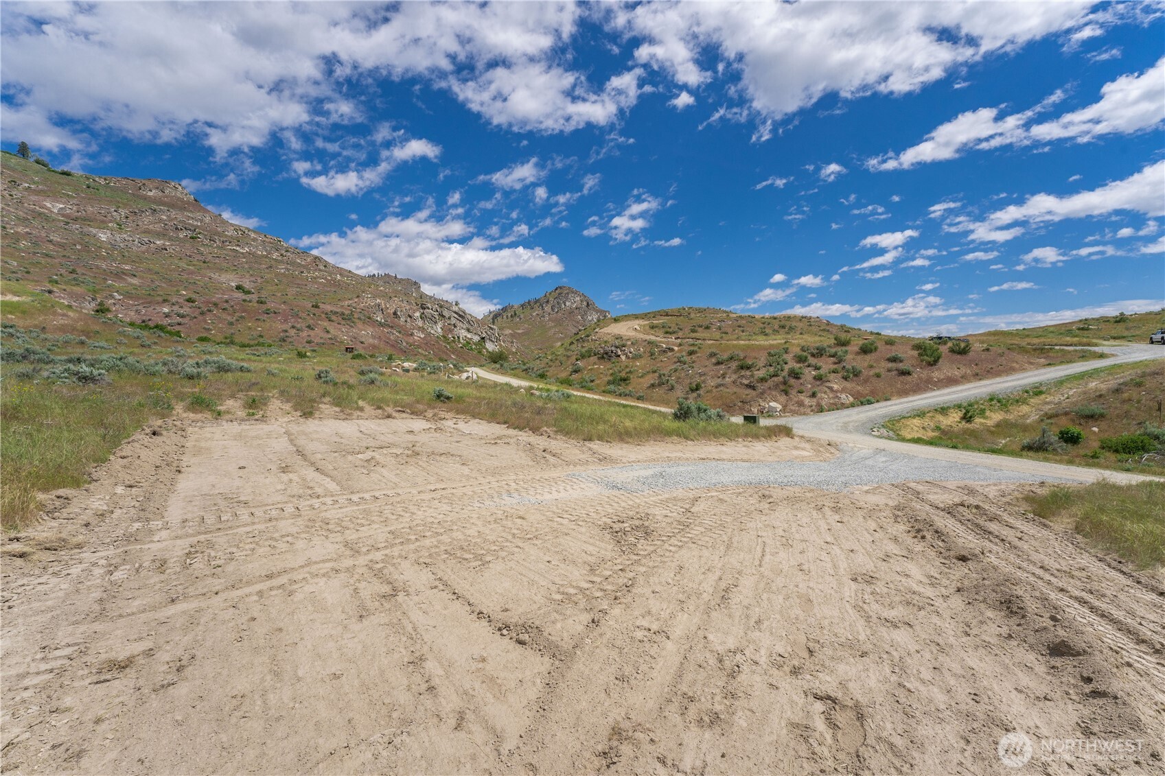 Vacant lot with a graded dirt clearing, gravel road, and dry rolling hills.