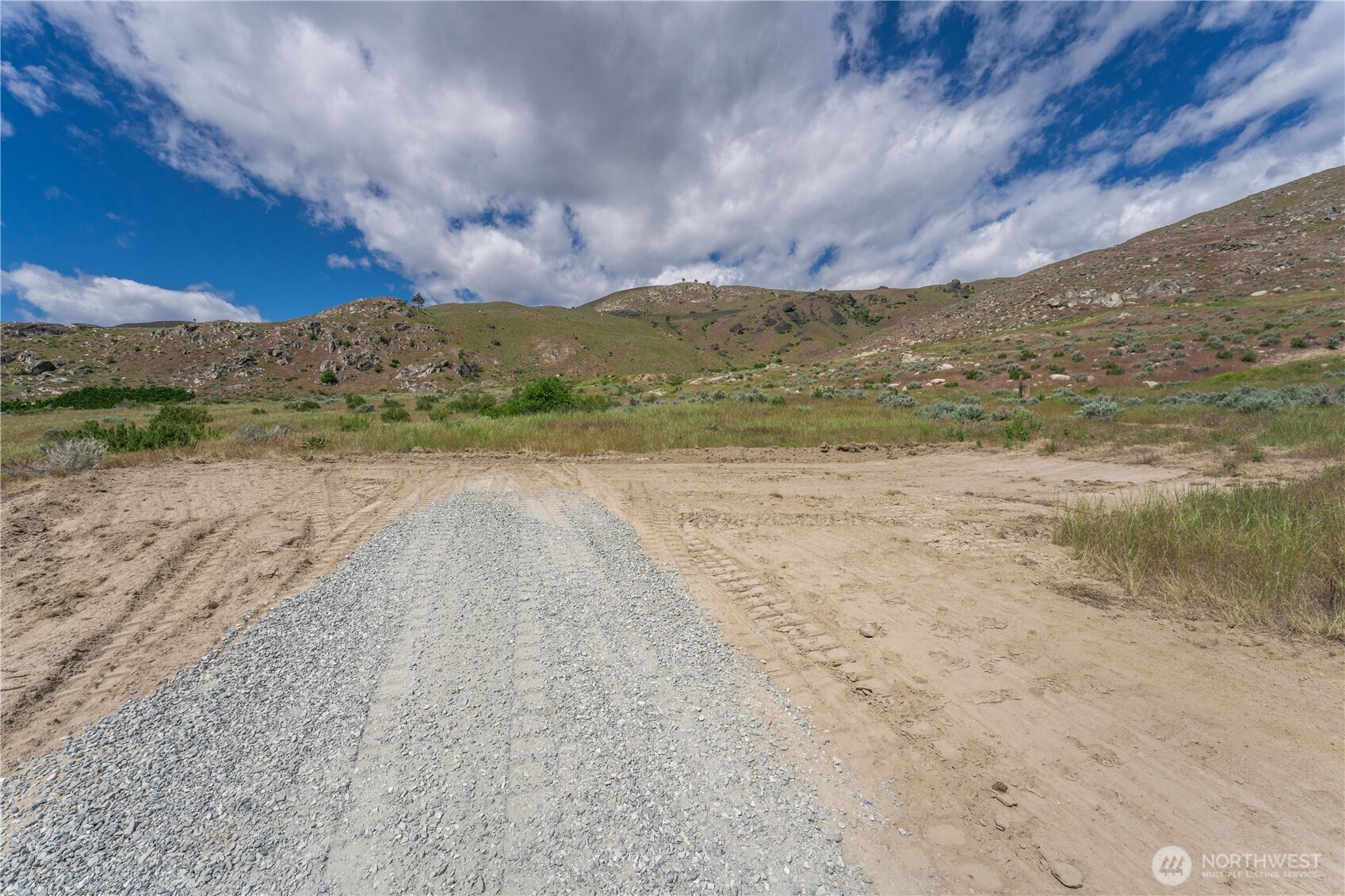 Vacant land with a gravel access track leading toward grassy, rocky hills.