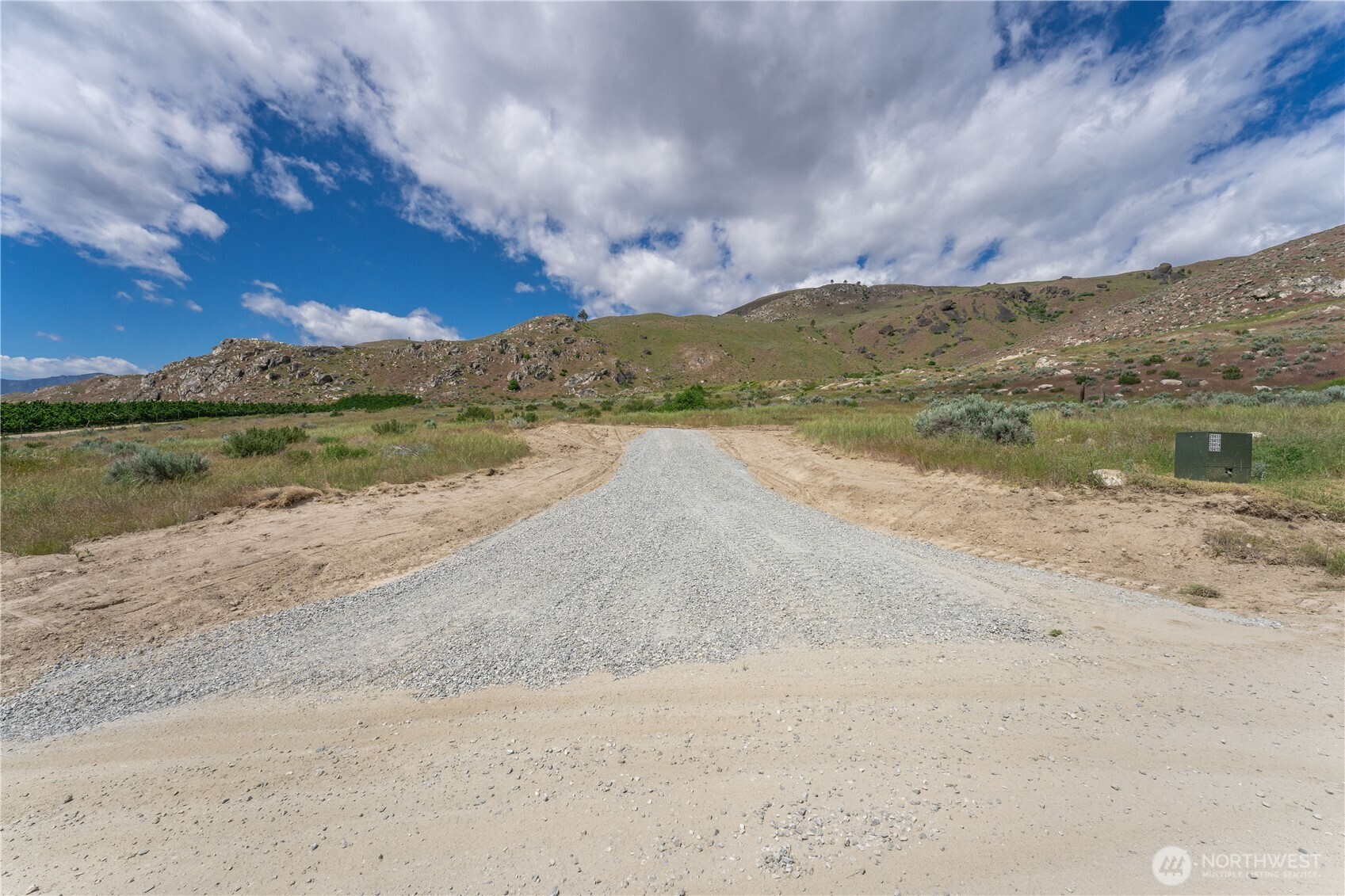 Gravel access road through open land toward rocky hills under a partly cloudy sky.