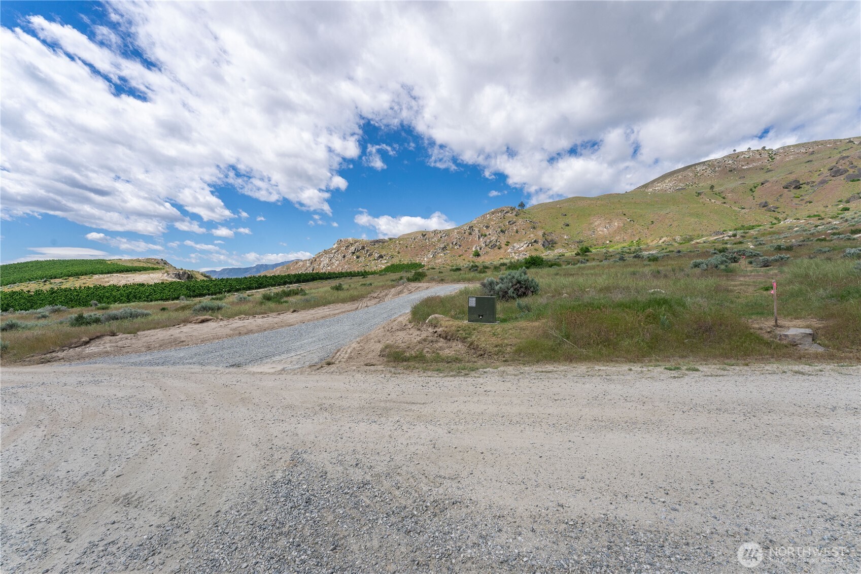 Undeveloped grassy property with a gravel access road, utility box, and rocky hills beyond.