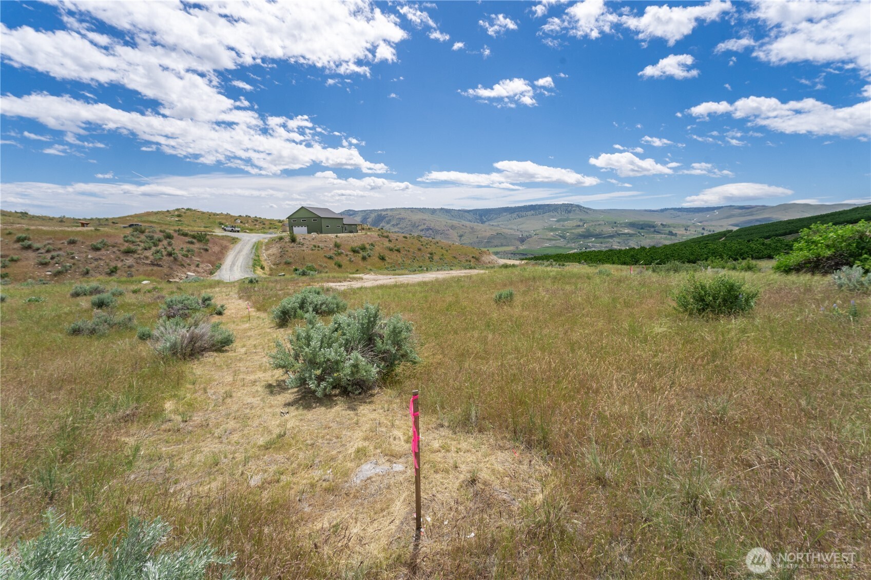 Grassy rural lot with survey markers, a gravel drive, a green outbuilding, and rolling hills beyond.