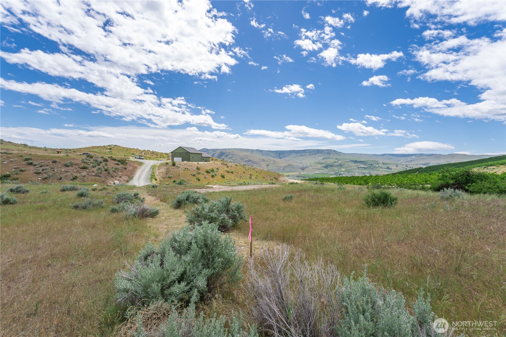 Open acreage with sagebrush, a dirt road, and a green outbuilding against rolling hills.