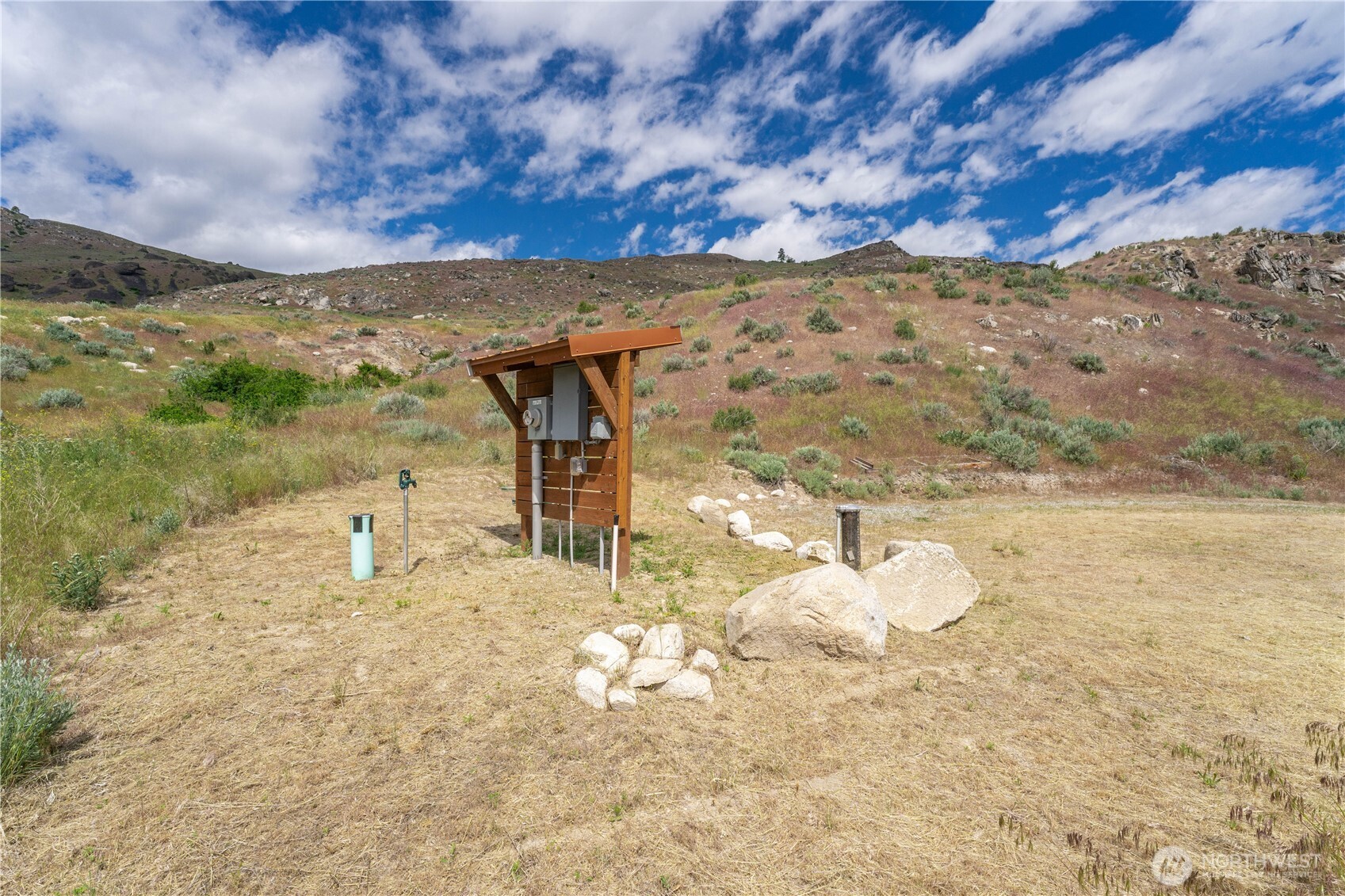 Open hillside lot with utility hookups and a wooden meter enclosure among dry grass and rocks.