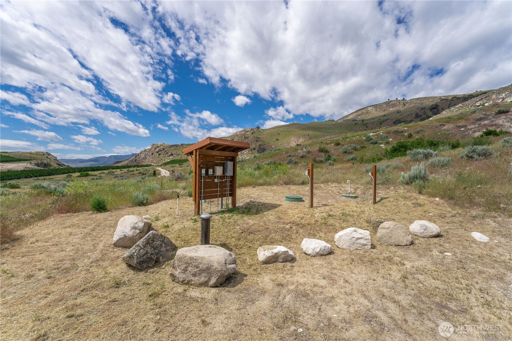 Open rural lot with a wooden utility shelter, scattered rocks, and hills in the distance.