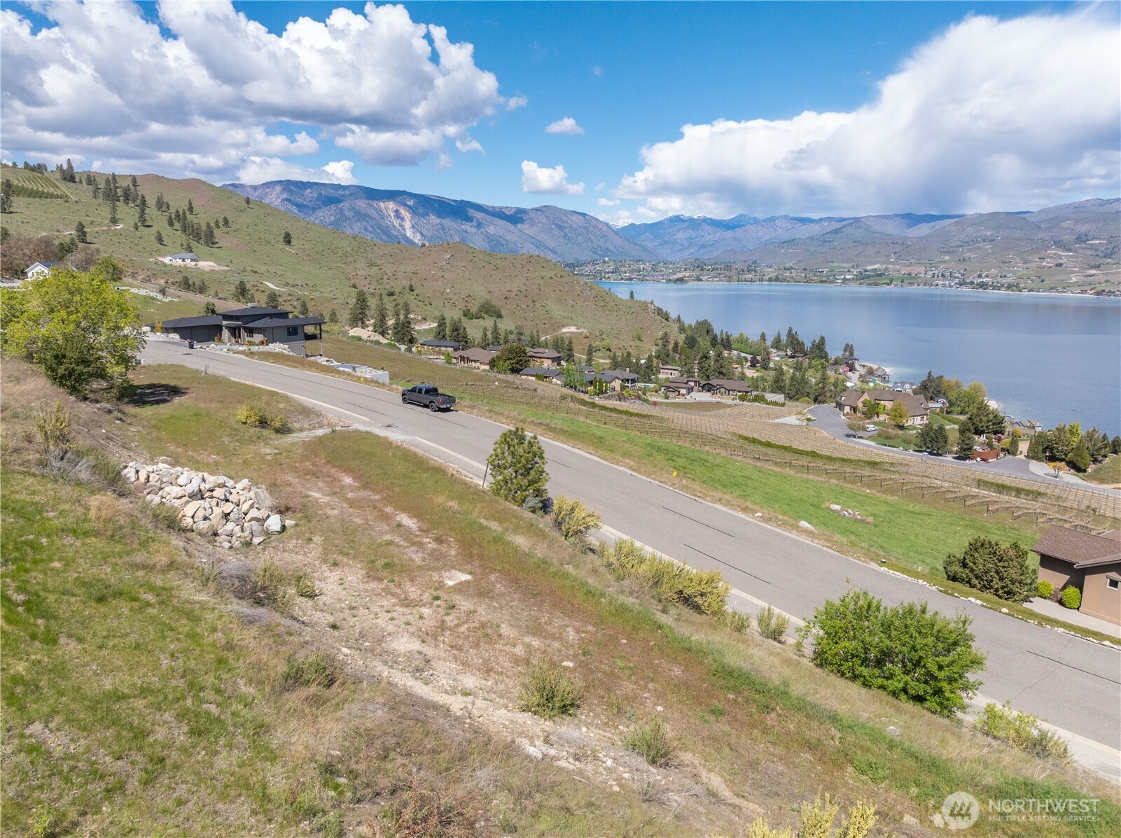 Hillside property beside a road overlooking Lake Chelan, with nearby homes, vineyard rows, and mountains.