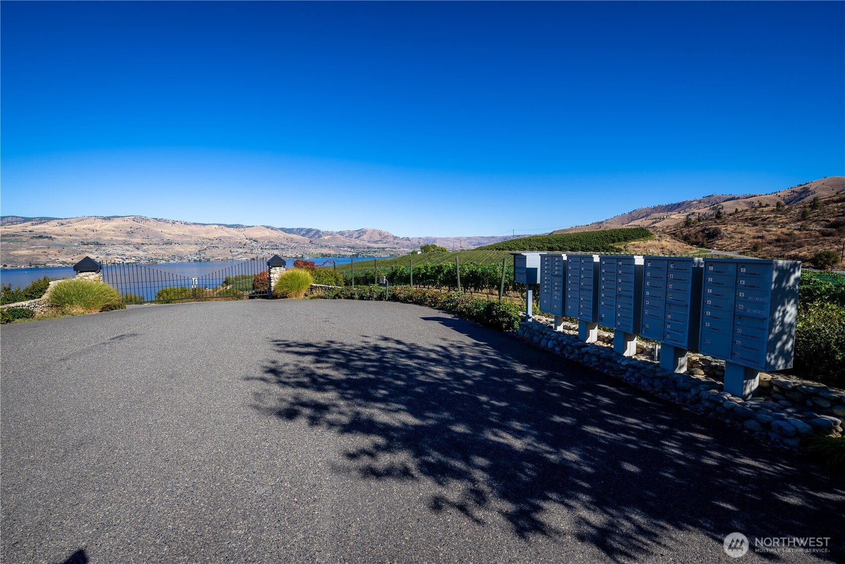 Community mailboxes beside a paved entrance, with vineyard, lake, and hills beyond.
