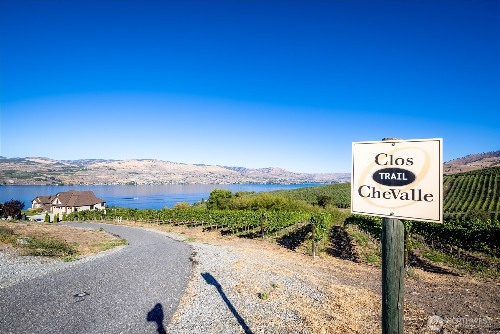 Clos Chevalle vineyard and trail sign overlook Lake Chelan, with a home and hills beyond.
