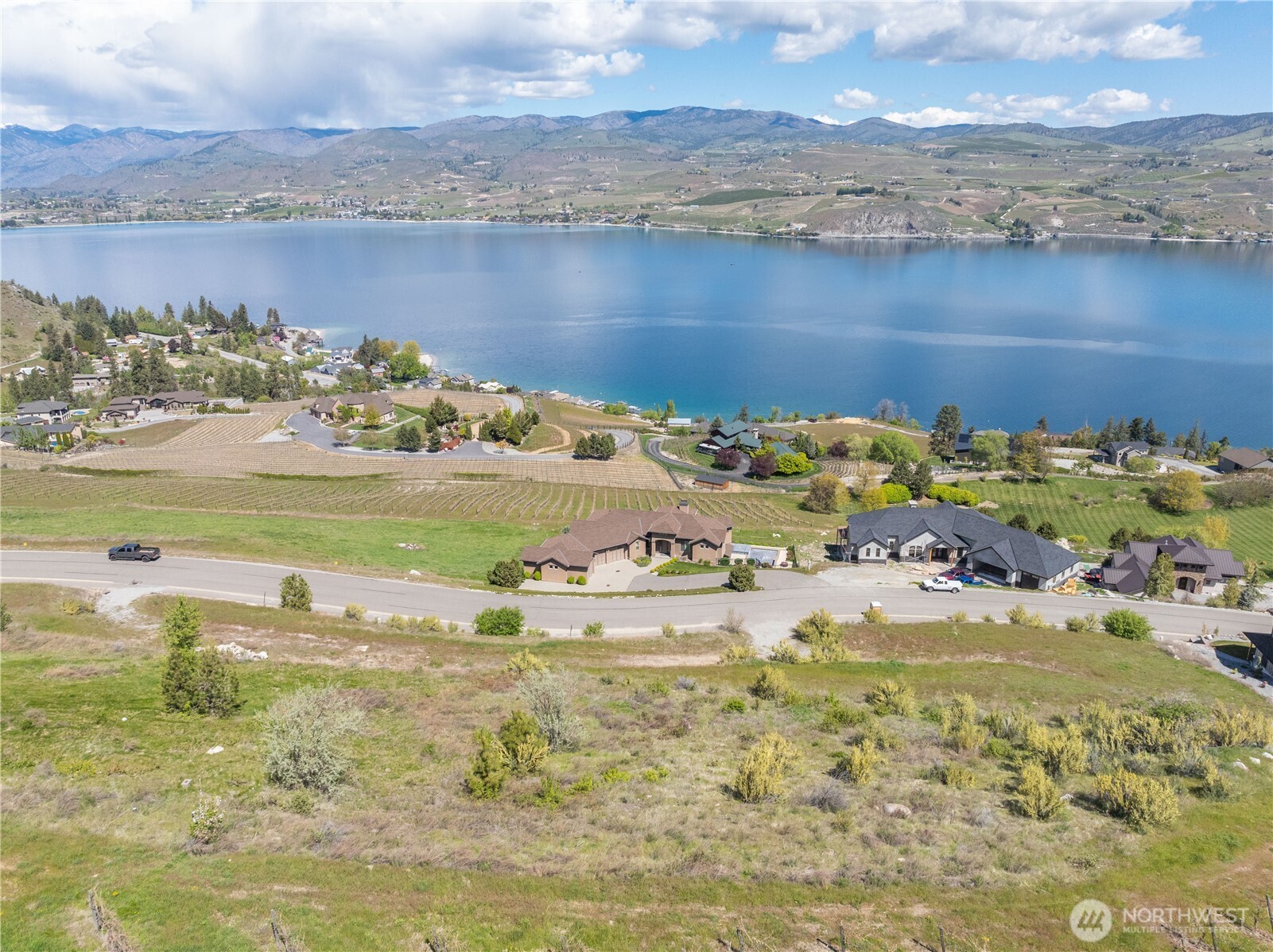 Open lakeside parcel beside vineyards, with homes, blue water, and mountains beyond.