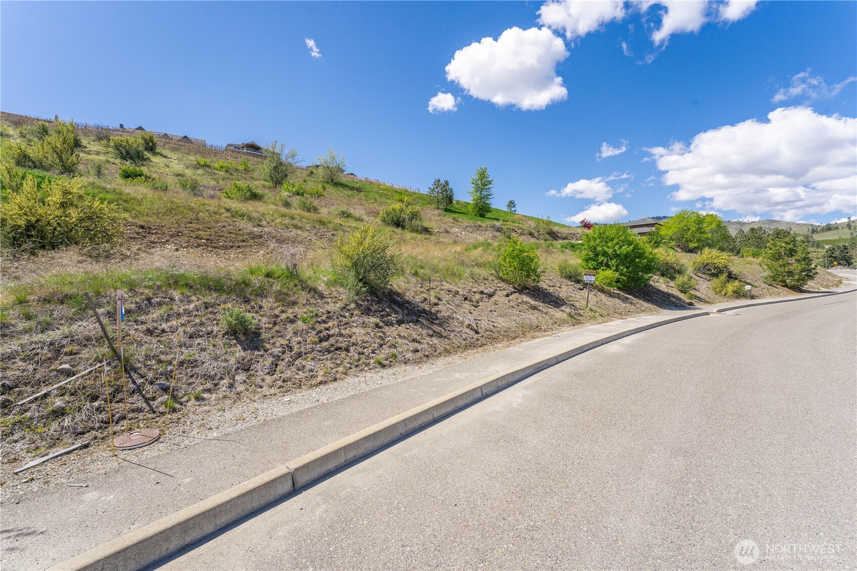 Vacant sloping lot beside a paved road, with sparse shrubs, utility markers, and a SOLD sign.