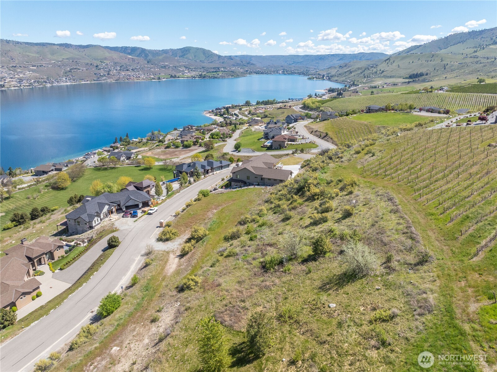 Aerial view of a hillside property overlooking Lake Chelan, with vineyard rows and homes nearby.