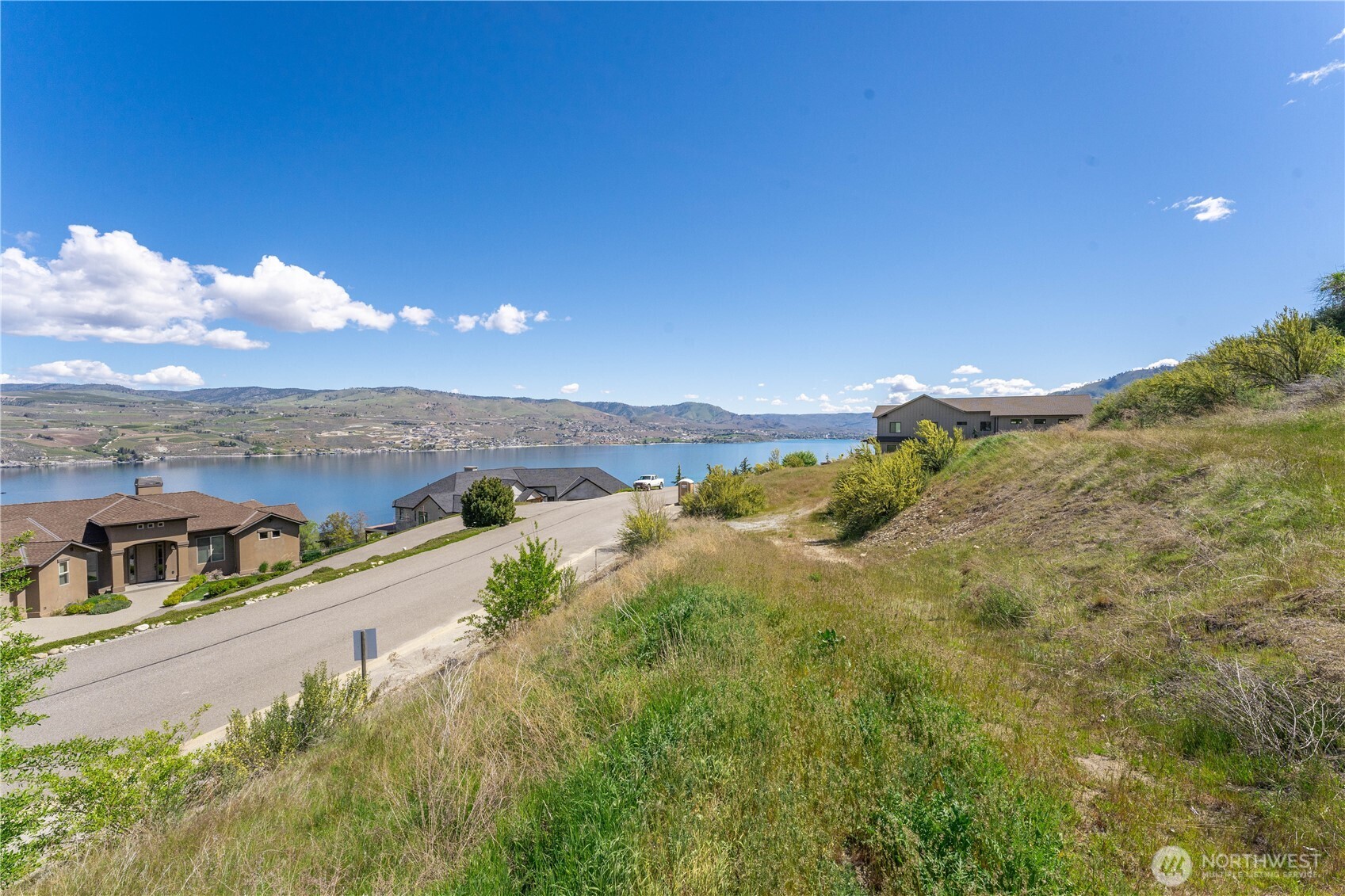 Hillside parcel overlooking Lake Chelan, with a sloping road, nearby homes, and distant hills.