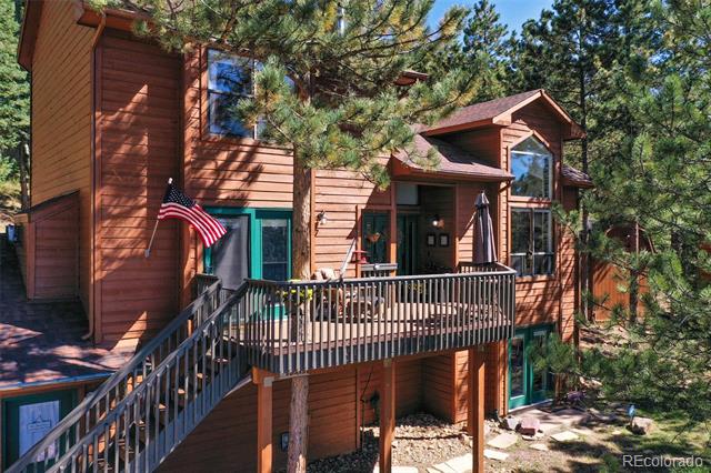 Wood-sided house with a raised deck, long staircase, American flag, and surrounding pine trees.