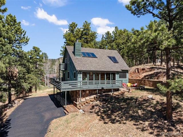 Blue-green house with a raised wraparound deck and solar panels, surrounded by pine trees.