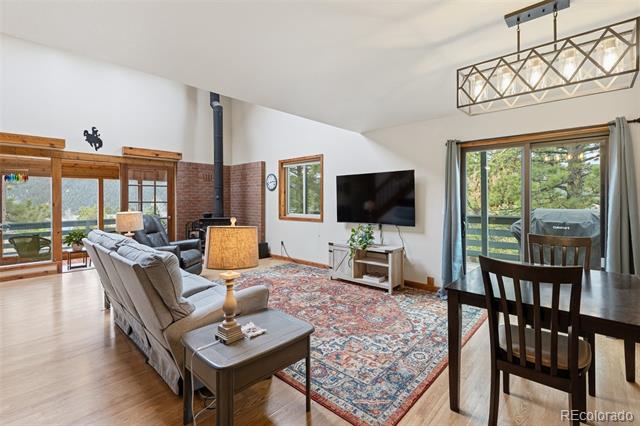 Living room with a gray sectional, brick fireplace, patterned rug, and large windows overlooking trees.