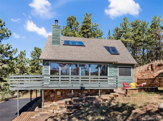 Green-sided home with a broad elevated deck, stone foundation, and roof windows among pine trees.