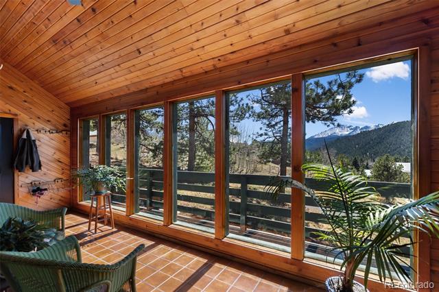 Wood-paneled sunroom with broad mountain-view windows, tiled floor, green chairs, and potted plants.