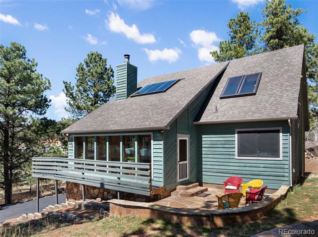 Teal-sided house with rooftop solar panels, a raised deck, and colorful patio chairs among pine trees.