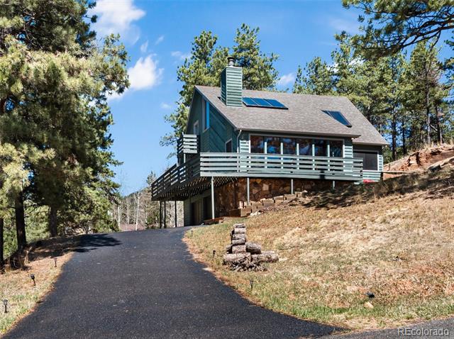 Teal mountain home with a wraparound deck, rooftop solar panels, and a paved driveway among pine trees.
