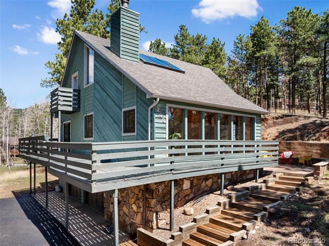 Teal mountain home with a raised wraparound deck, stone foundation, and rooftop solar panels.