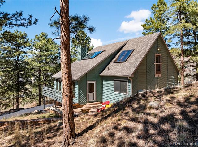 Green A-frame house on a wooded hillside, with a raised deck, skylights, and rooftop solar panels.