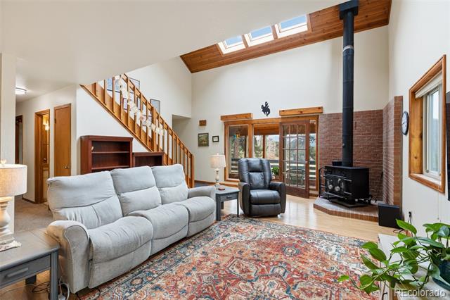 Living room with vaulted skylit ceiling, wooden staircase, brick fireplace, and patterned rug.