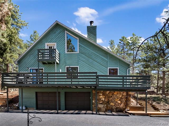 Teal gabled house with a long deck, two garage doors, and stonework among pine trees.