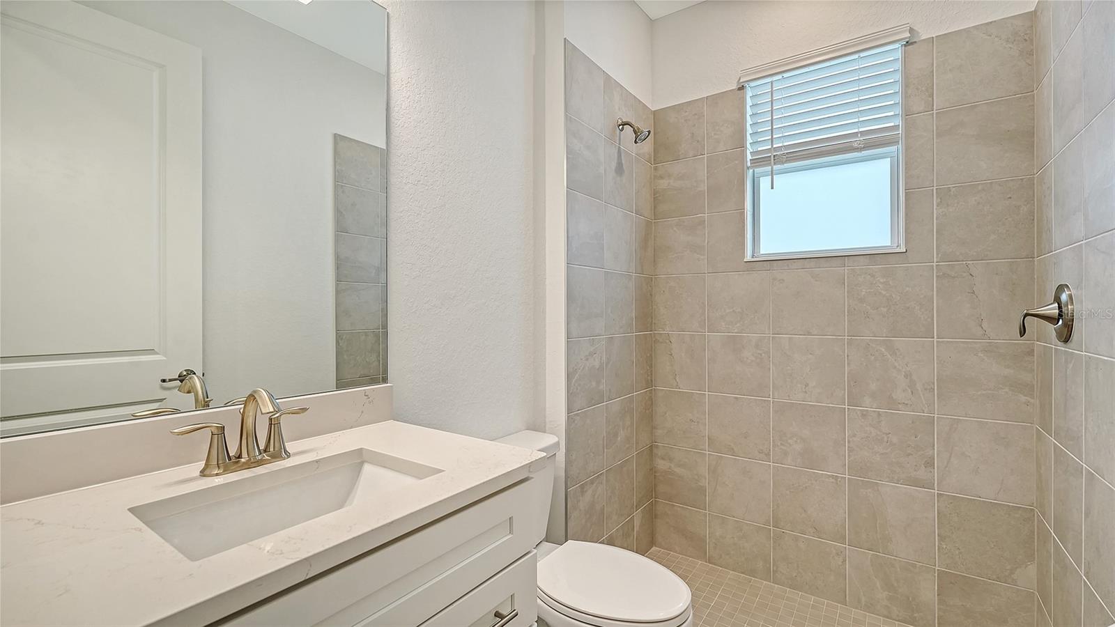 Bathroom with a white vanity and beige-tiled shower featuring a window with blinds.