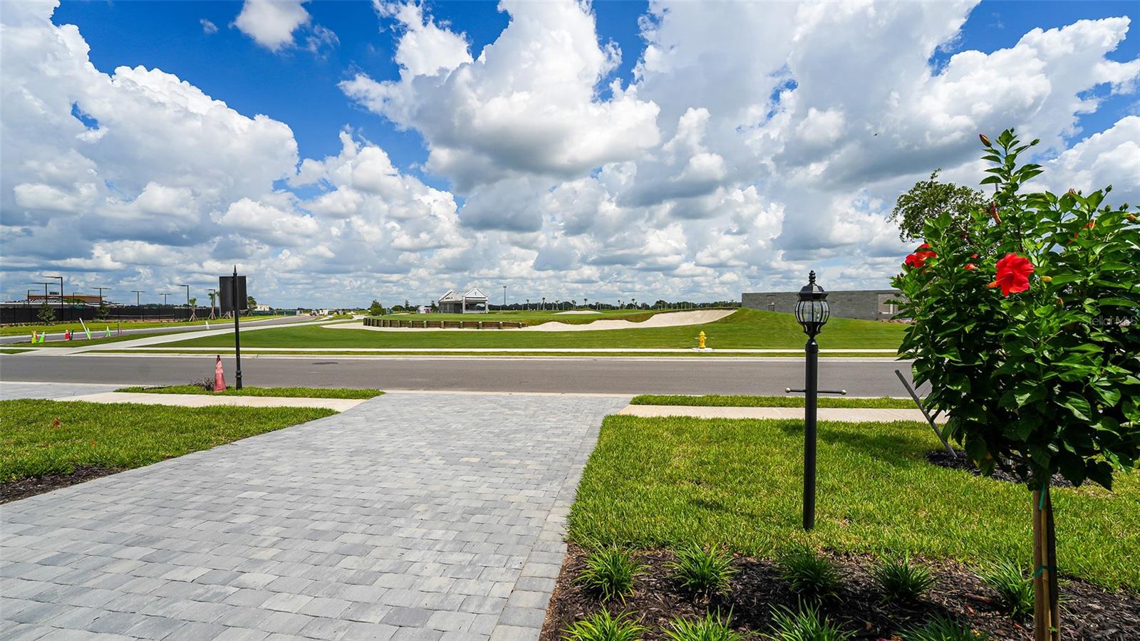 Paved driveway and landscaped lawn beside a street, with a pavilion and grassy mounds across the road.