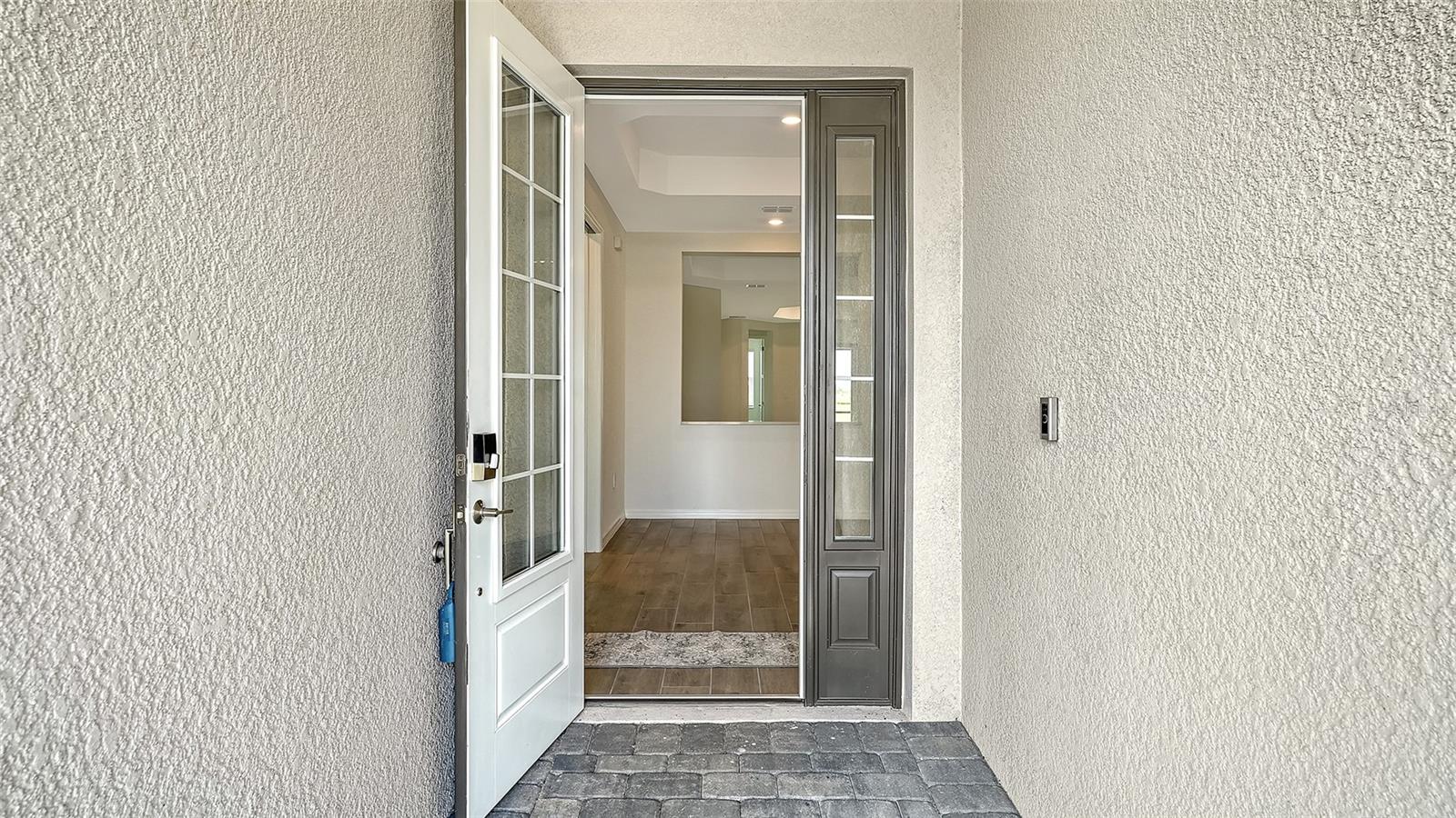 Open glass-panel front door reveals a tiled foyer and hallway from the covered paver porch.