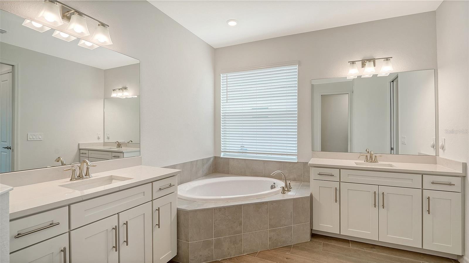 Bathroom with dual white vanities and a corner soaking tub beneath a window.