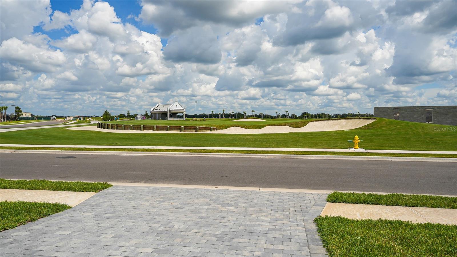 Golf course with long sand bunker and clubhouse across the street from a paved driveway.