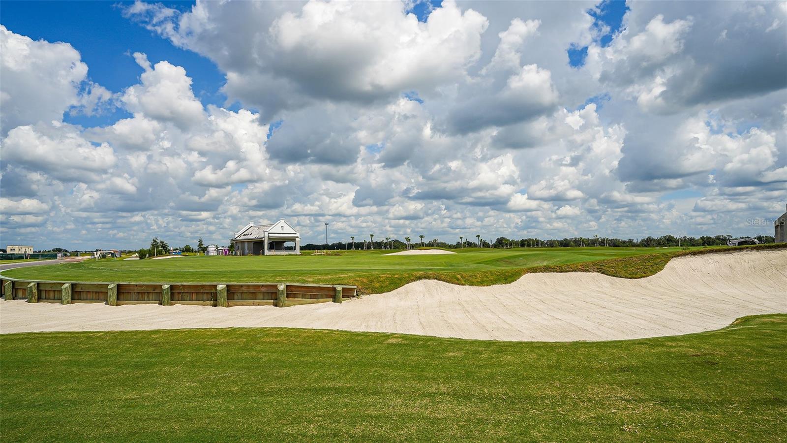 Panoramic golf course with a wide sand bunker and small clubhouse beneath towering clouds.
