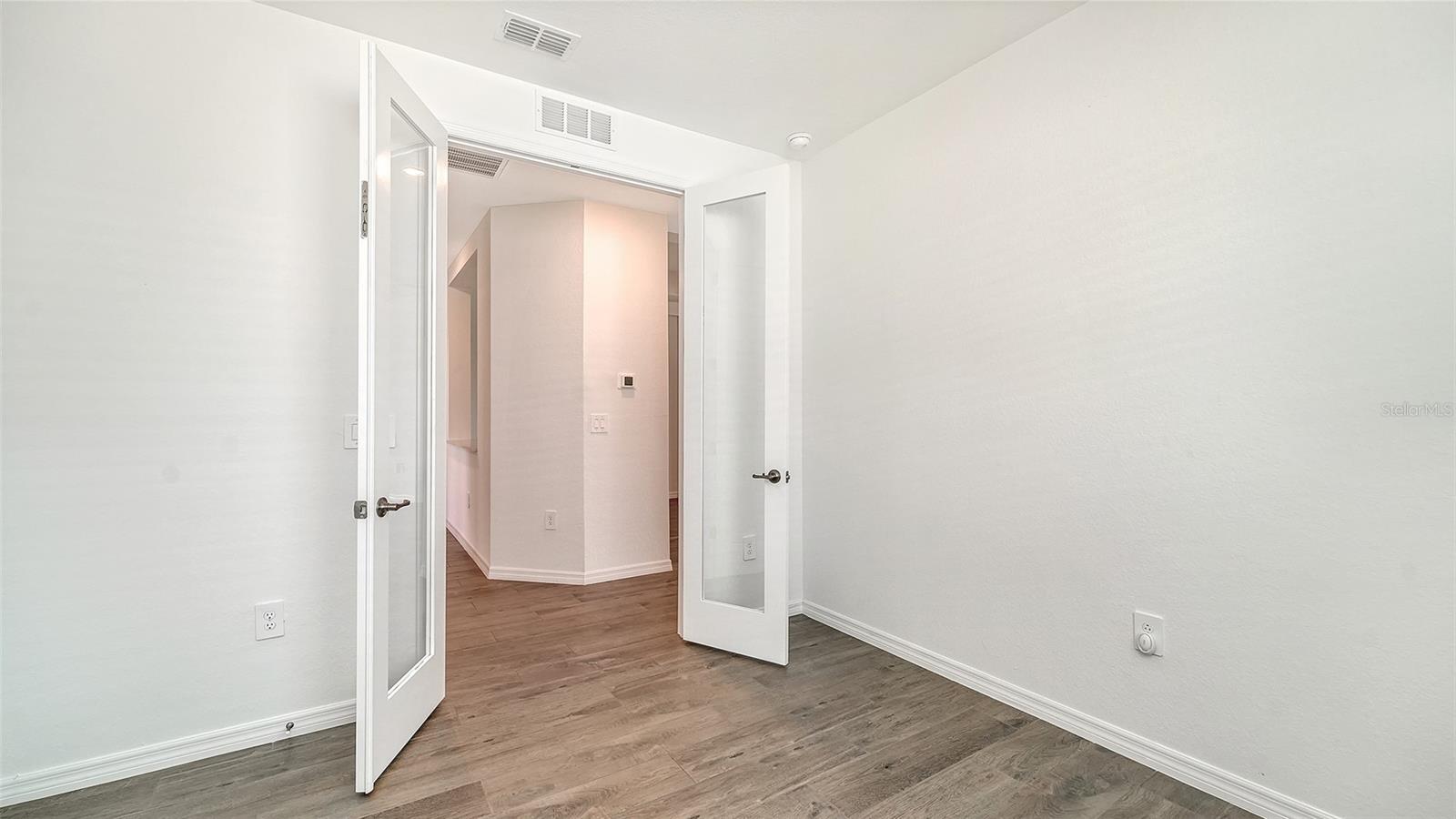 Empty room with wood-look flooring and open glass-paneled doors to a hallway.