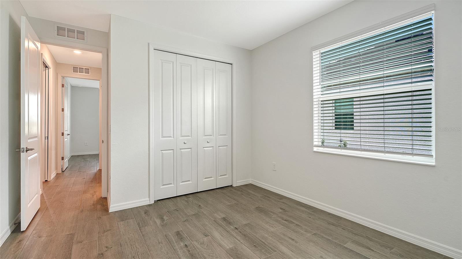 Empty bedroom with a four-panel closet, a wide window with blinds, and gray wood-look tile flooring.