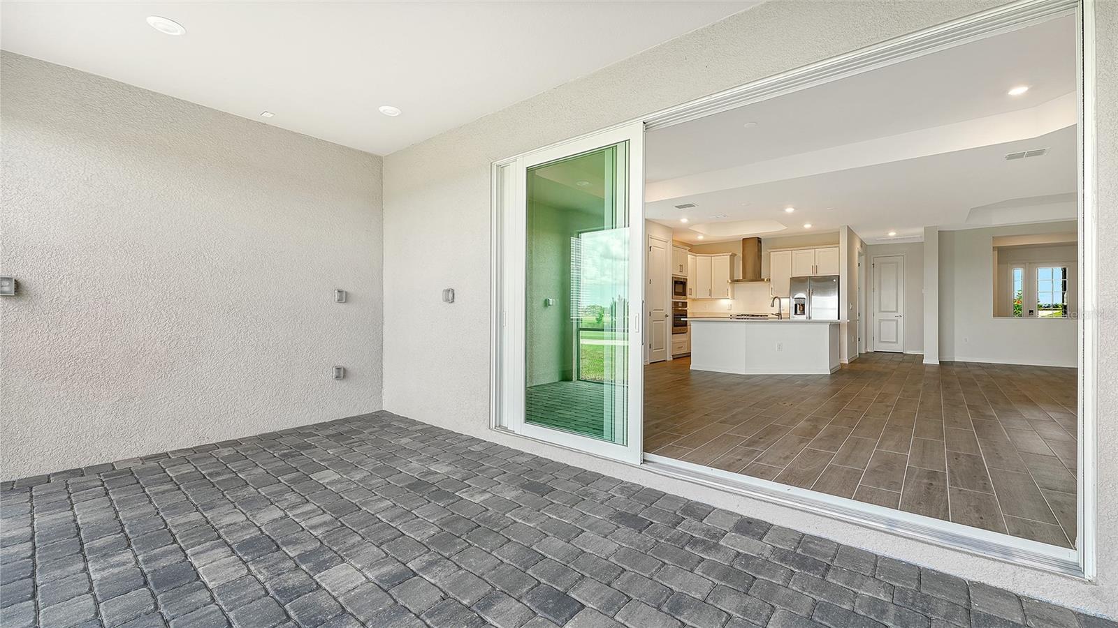 Covered paver lanai opening through sliding glass doors to a bright kitchen with an island.