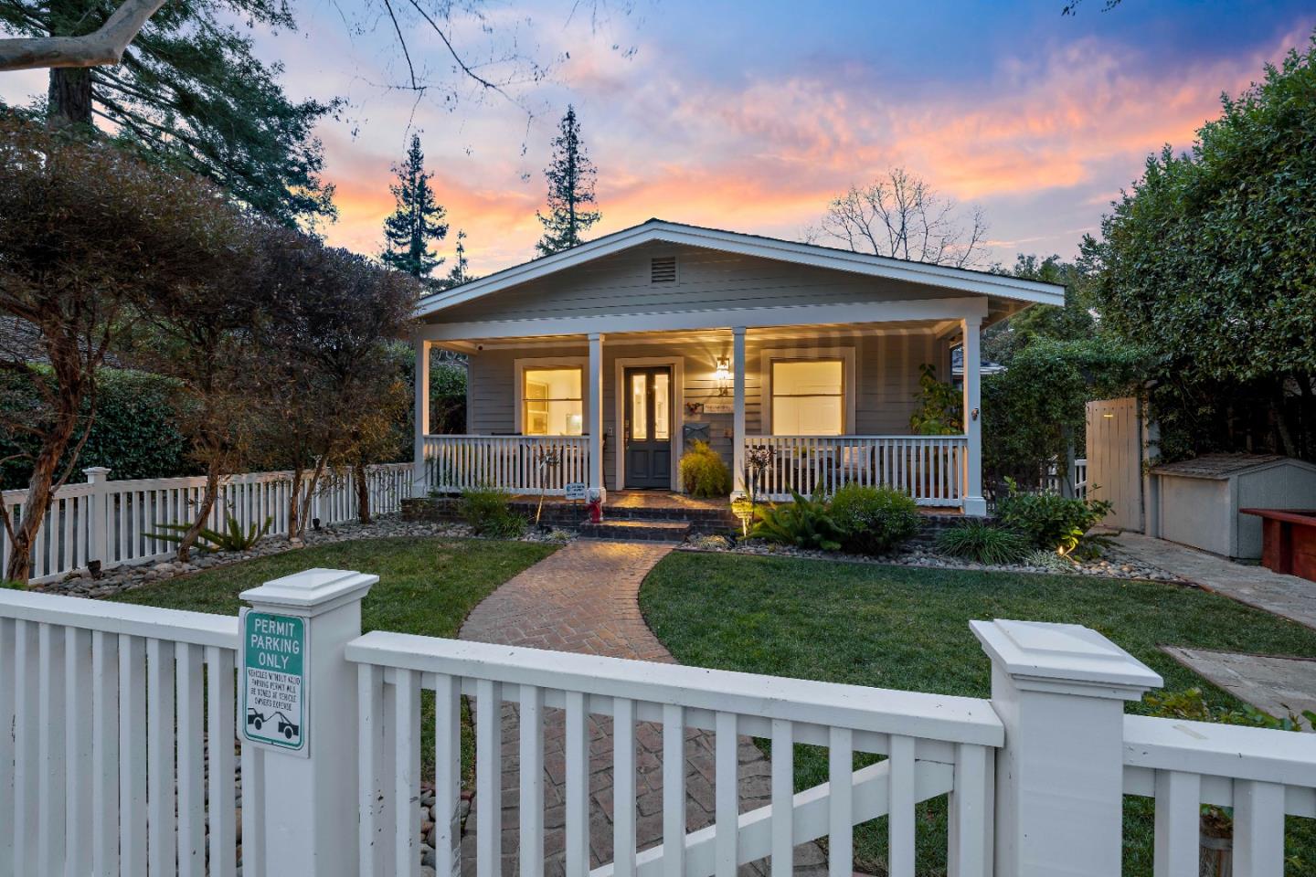 Gray home with a covered porch and curved brick walkway behind a white fence at dusk.
