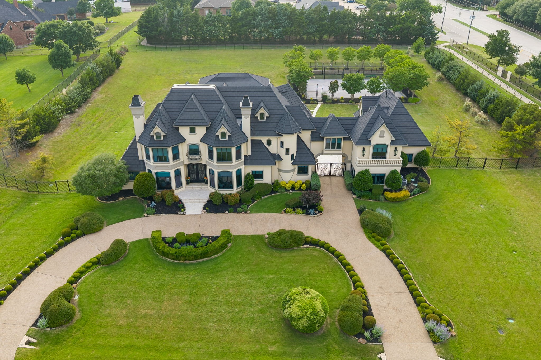 Aerial view of a cream mansion with dark tiled roofs, formal landscaping, and a circular driveway.