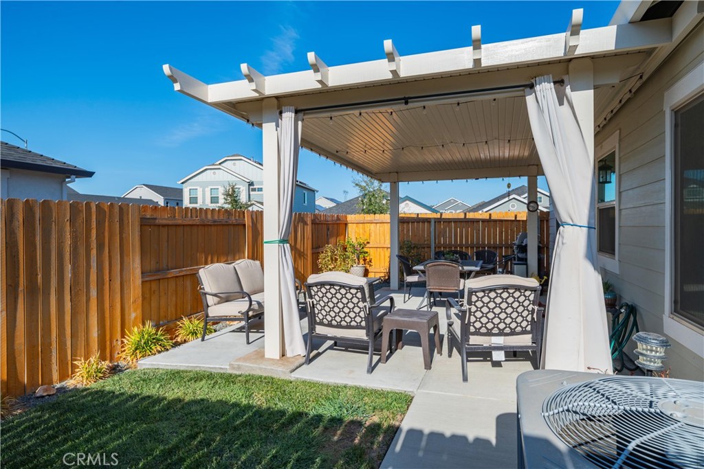 Backyard patio with a curtained pergola, outdoor seating, wooden fence, and small lawn.