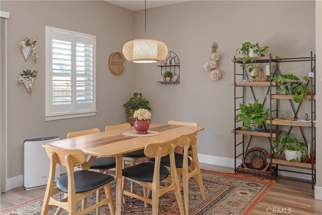 Dining room with a light wood table, patterned rug, and plant-filled shelving.