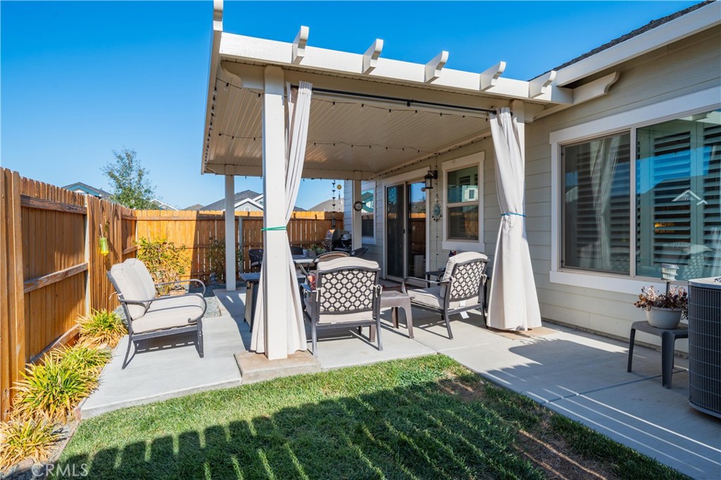 Covered backyard patio with a pergola, curtains, and outdoor seating beside a fenced lawn.