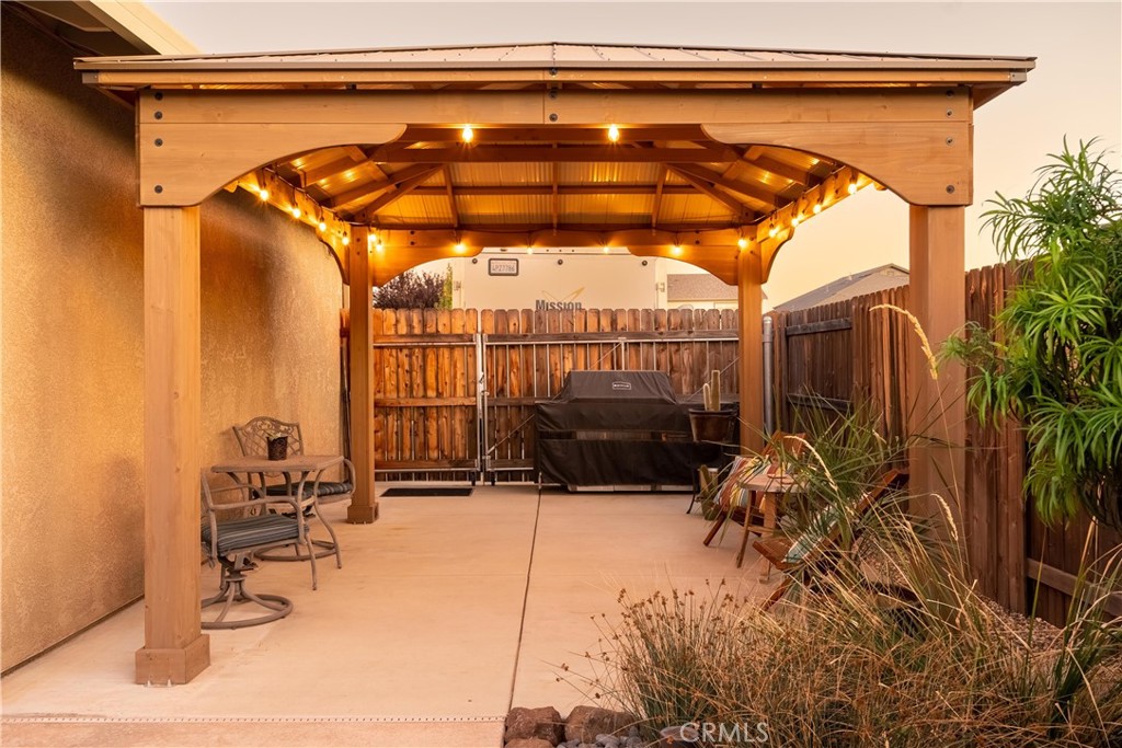 Covered patio beneath a wooden pergola with warm lights, outdoor seating, and a barbecue grill.