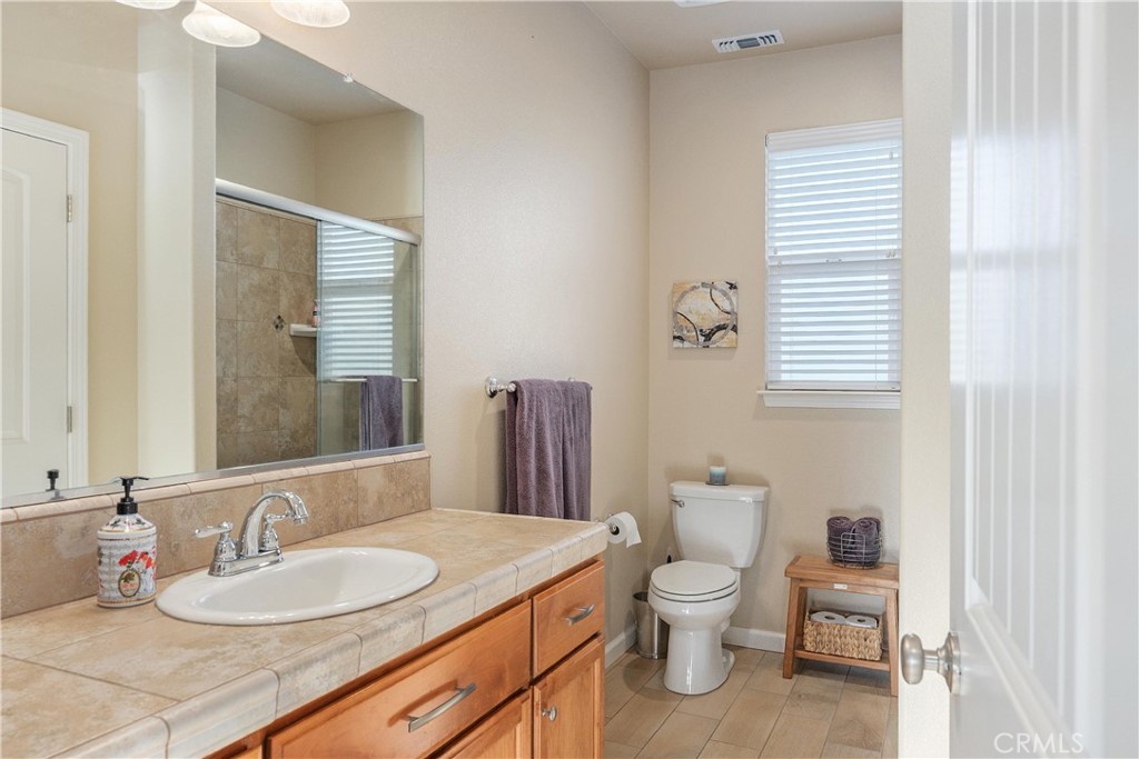 Bathroom with tan tiled vanity, wood cabinets, glass shower, and window with blinds.