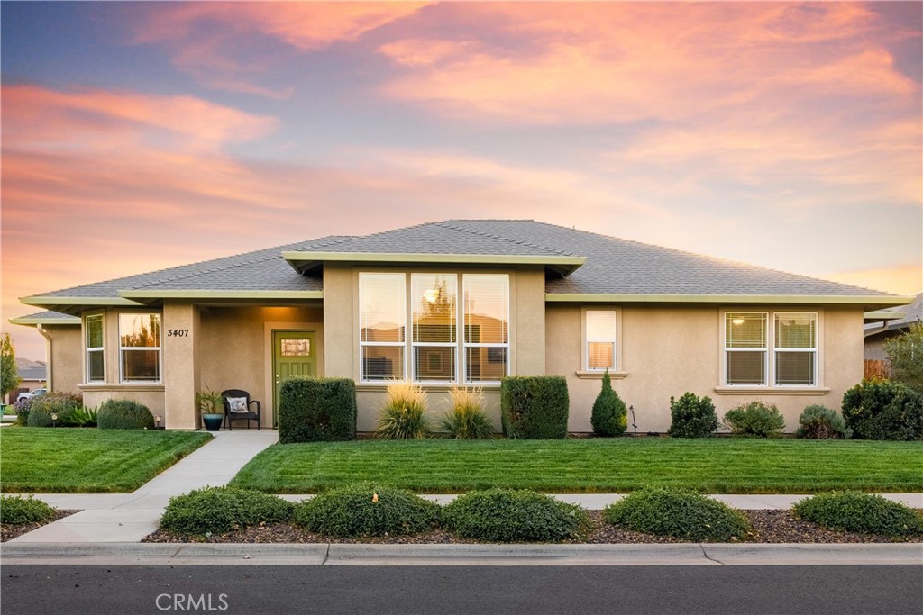 Single-story beige stucco home with a gray roof, landscaped lawn, and pink sunset sky.